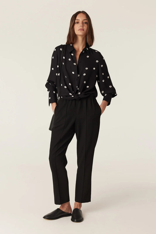 Woman wearing the Cable Melbourne Hepburn Blouse in Black Spot, tied at the hem over black trousers